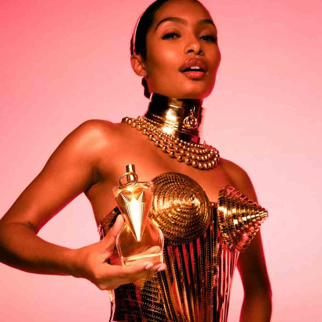 Woman in a gold corset holds Jean Paul Gaultier perfume bottle, both reflecting a bold and glamorous metallic design.