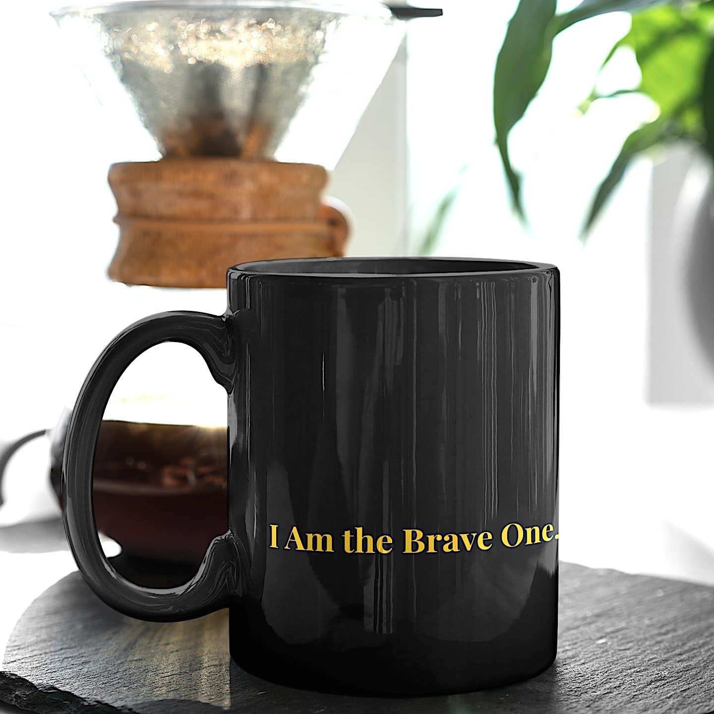 Black ceramic mug with gold "I Am the Brave One" text, placed beside a coffee brewer on a dark surface with sunlight in the background.