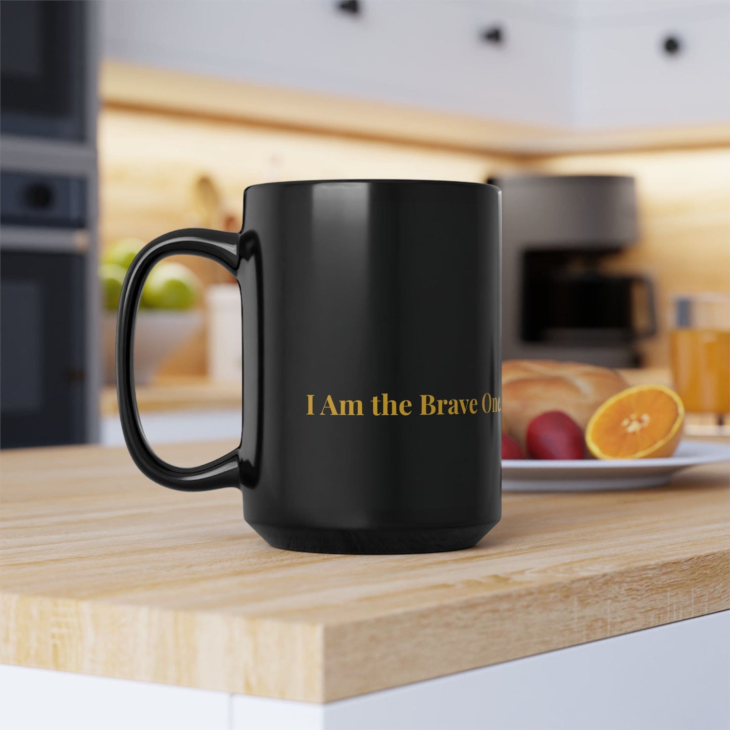 Black ceramic mug with gold "I Am the Brave One" text, placed on a wooden kitchen counter with breakfast in the background.