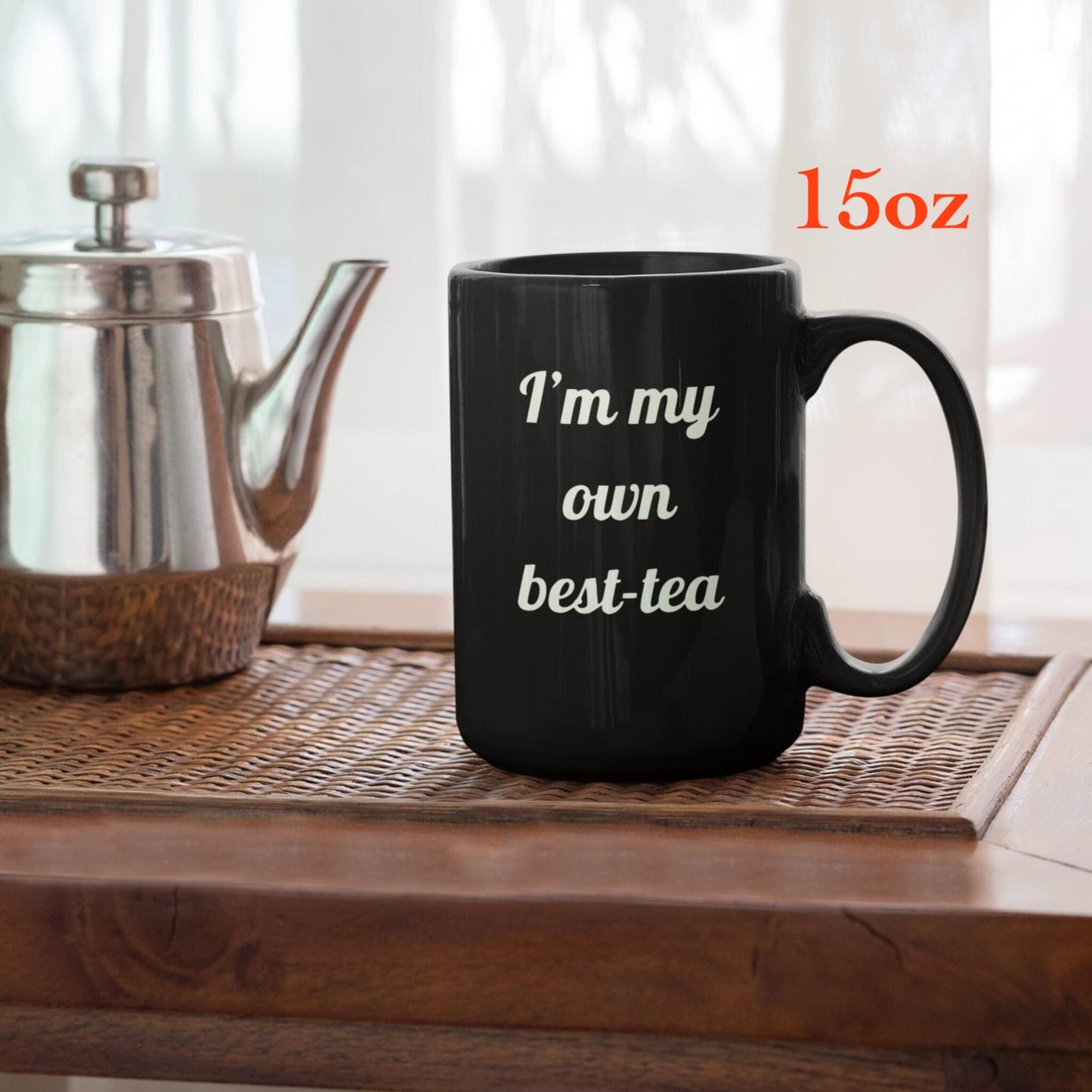 Black ceramic mug with “I’m my own best-tea” quote on wooden table beside a silver teapot.