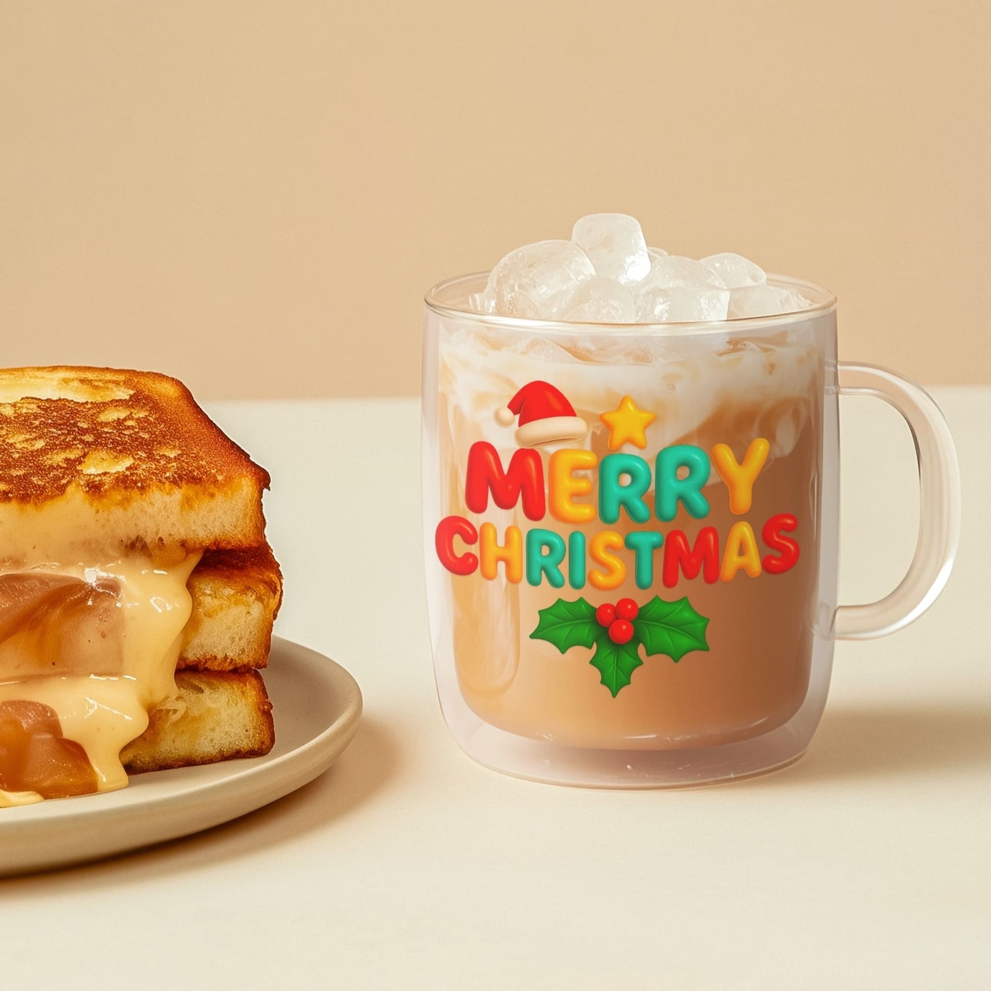 Festive clear glass mug with iced latte and Merry Christmas design next to a grilled cheese sandwich on a beige table.
