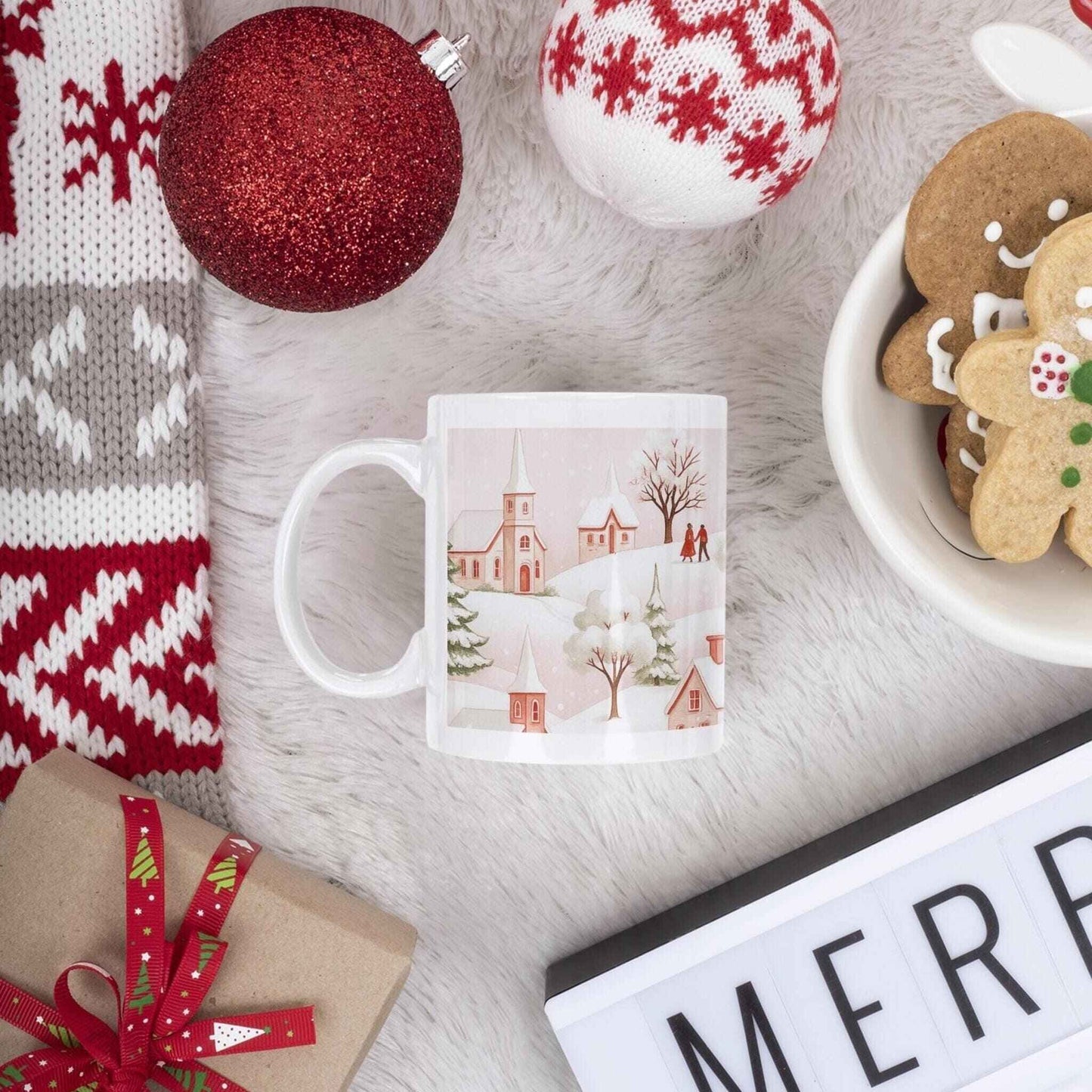 White ceramic mug with winter village design surrounded by Christmas decorations, gingerbread cookies, and cozy holiday items on fluffy white surface