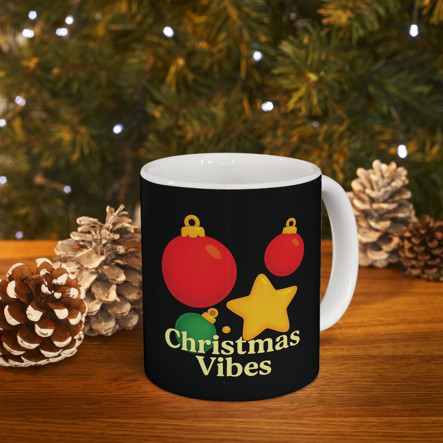 Black ceramic mug with red, green, yellow ornaments and “Christmas Vibes” text, on wood table with pinecones and tree lights.