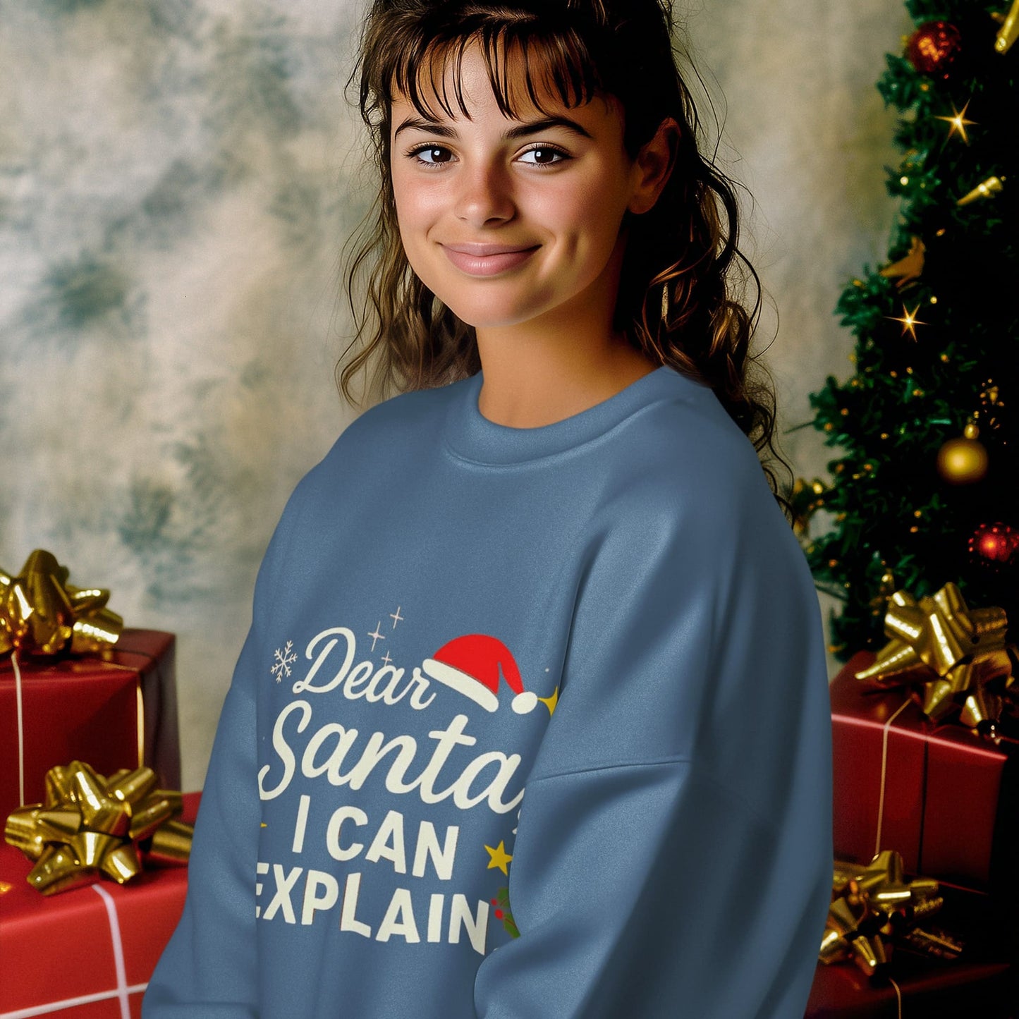 Young woman smiling in blue Christmas sweatshirt with “Dear Santa I Can Explain” text, sitting by gifts and holiday tree.