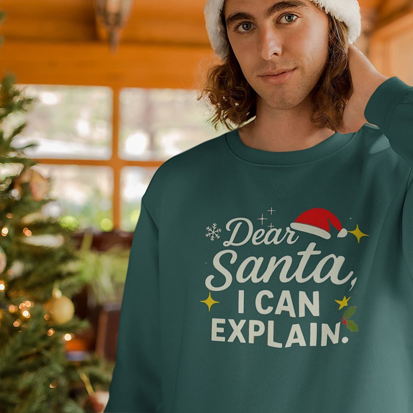 Man in green Christmas sweatshirt with “Dear Santa I Can Explain” text, wearing Santa hat by decorated holiday tree indoors.