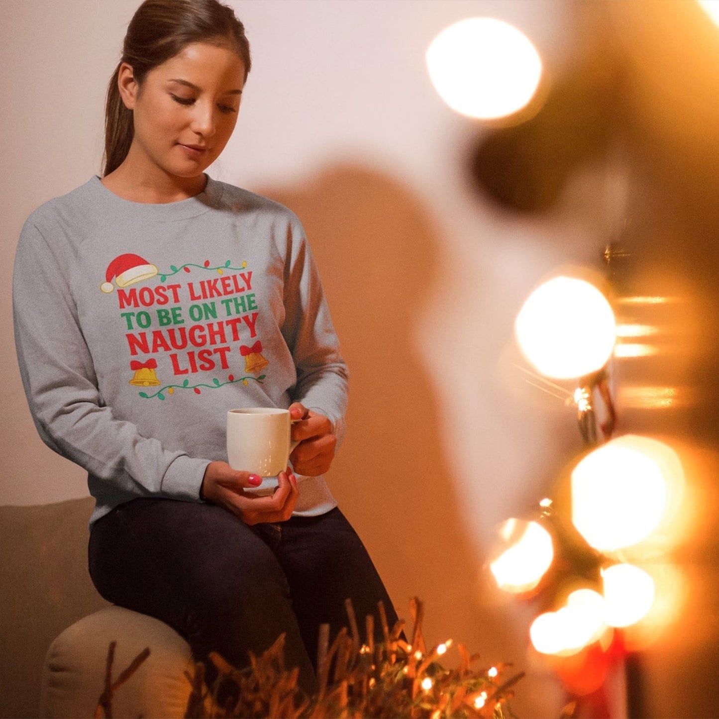 Woman wearing a Gildan 18000 Sport Gray Christmas sweatshirt with the text “Most Likely to Be on the Naughty List,” sitting indoors holding a mug by holiday lights.