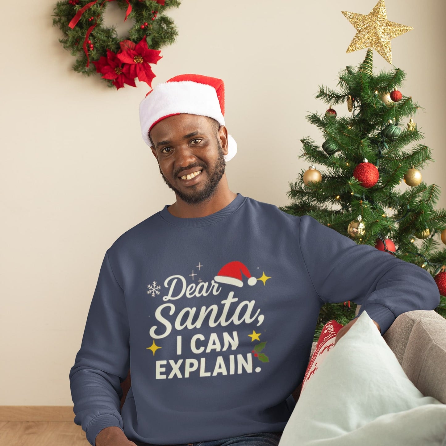 Smiling man in a Santa hat sitting by a decorated Christmas tree, wearing a navy sweatshirt with the text “Dear Santa, I Can Explain.”