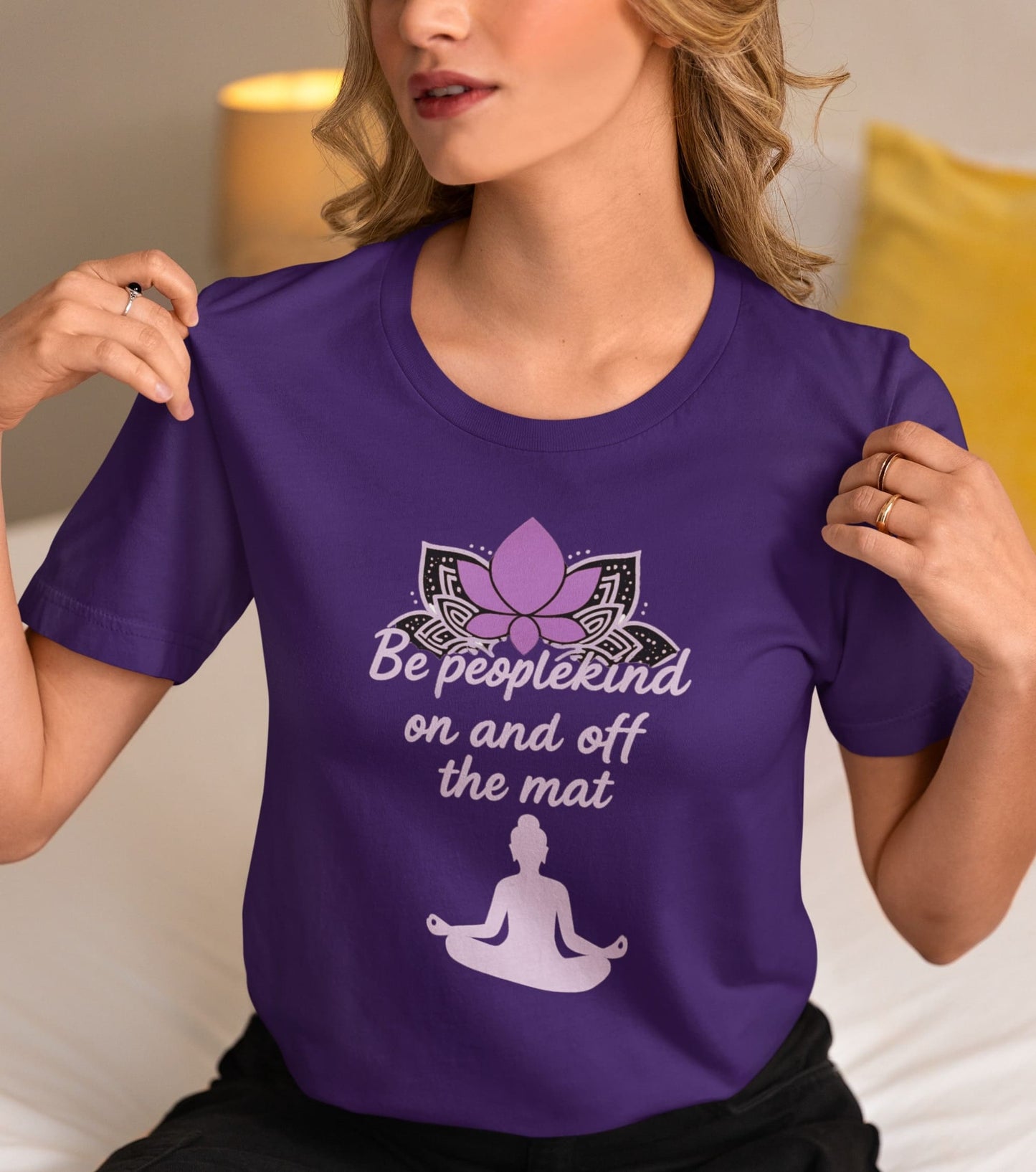 Woman wearing Team Purple Bella Canvas 3001 shirt with “Be peoplekind” yoga design, sitting on bed and holding the sleeves.