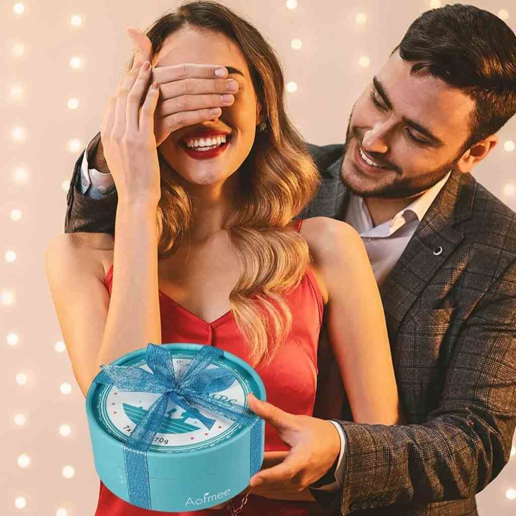 A man surprises a smiling woman with a blue gift-wrapped bath bomb set, creating a romantic vibe with soft background lights.
