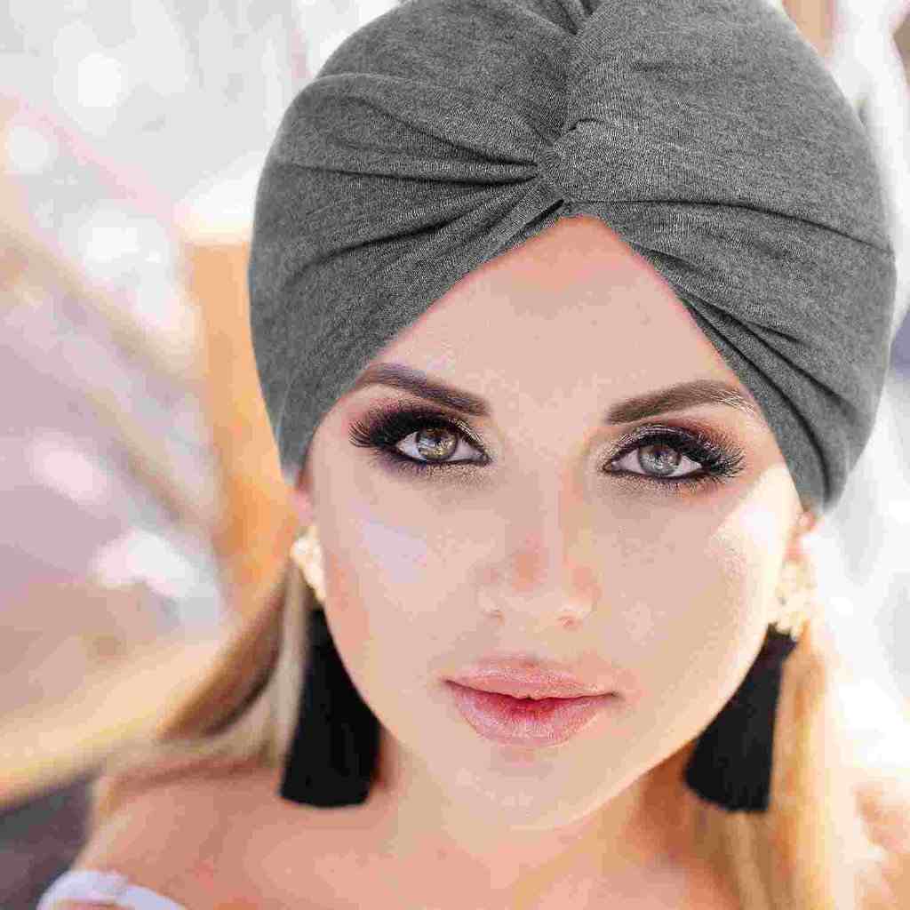 Close-up of a woman with striking eye makeup, wearing a gray turban and black tassel earrings, looking directly at the camera.