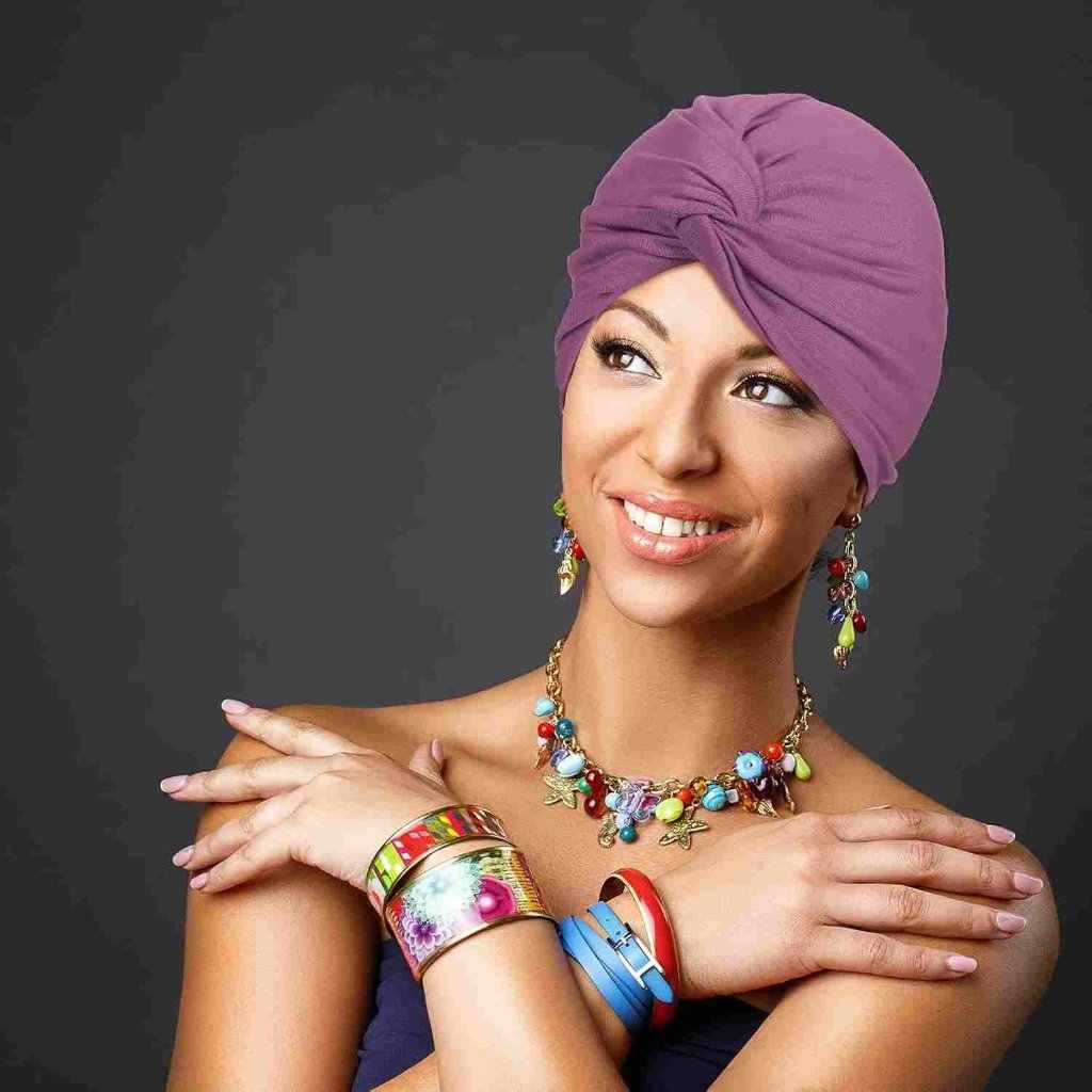 Smiling woman in a purple turban, adorned with a colorful necklace, matching earrings, and vibrant bangles on her arms.