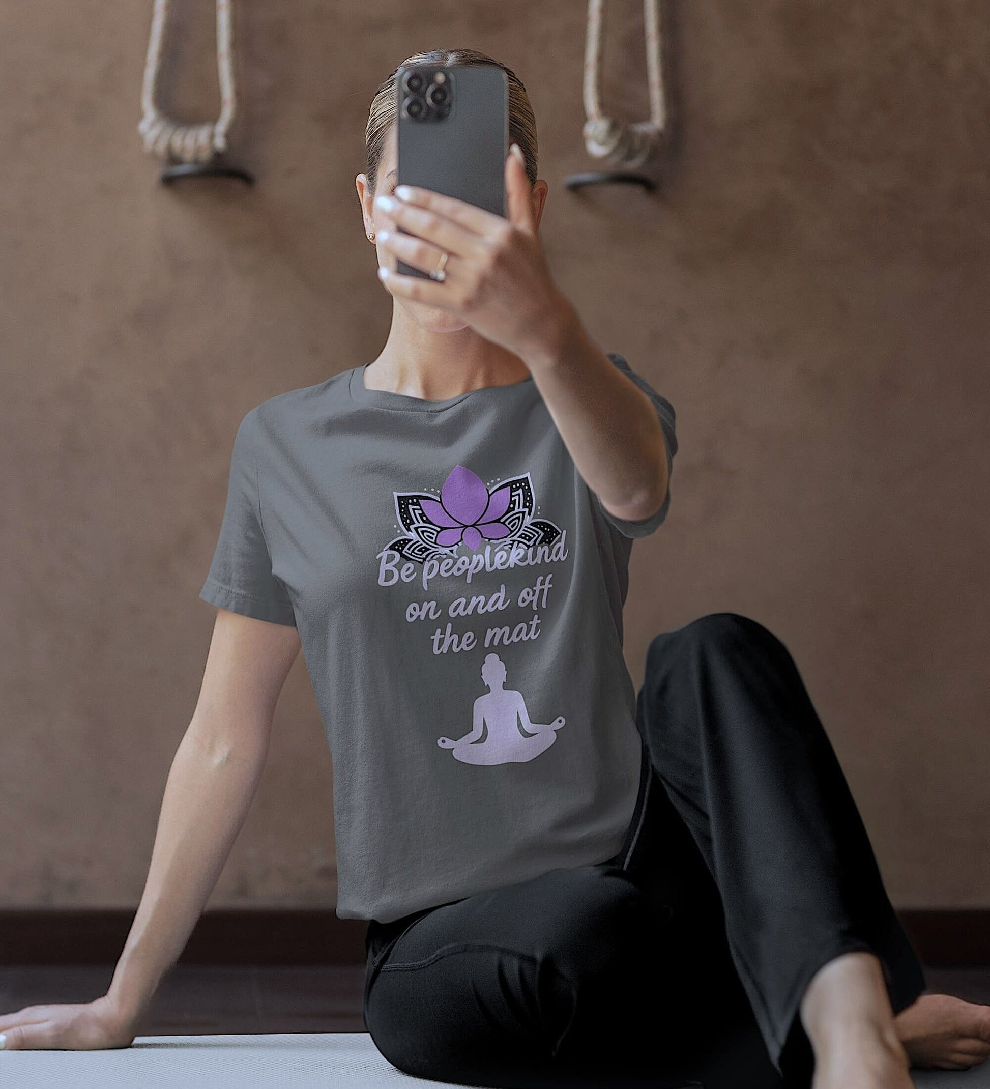 Woman wearing Asphalt Bella Canvas 3001 t-shirt with lotus and "Be peoplekind" quote, taking a selfie after yoga on mat.
