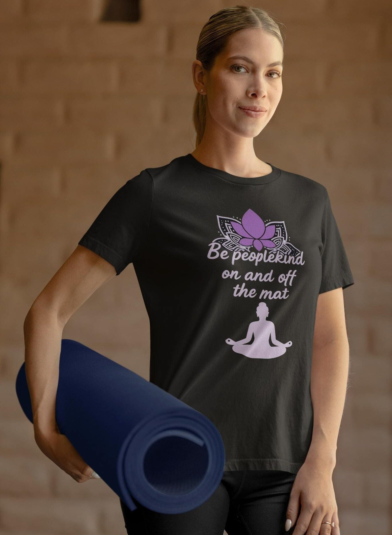 Woman wearing black Bella Canvas 3001 t-shirt with lotus and "Be peoplekind" quote, holding a yoga mat before class.