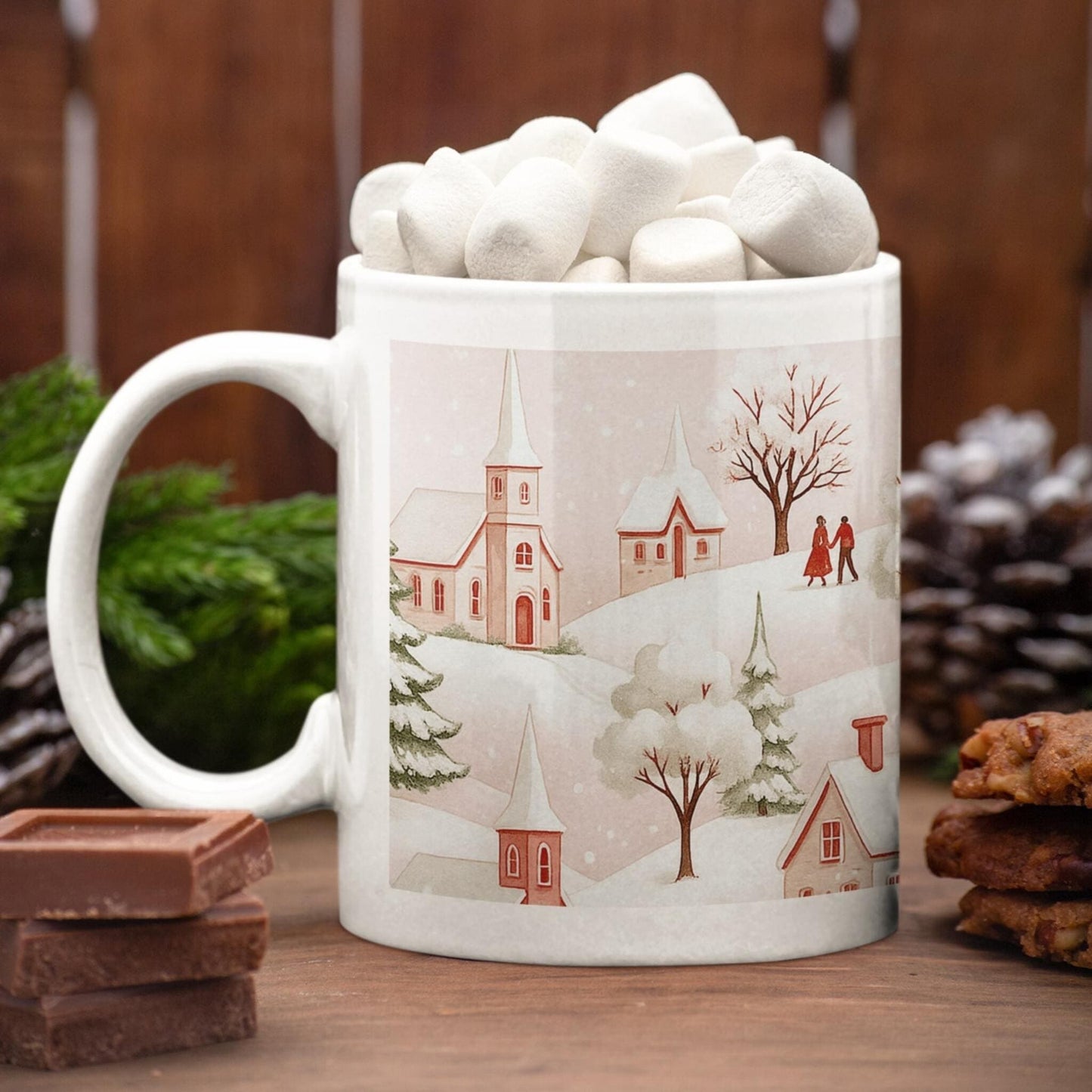 White coffee mug with winter village illustration filled with marshmallows, surrounded by pinecones, chocolate, cookies, and evergreen holiday decor
