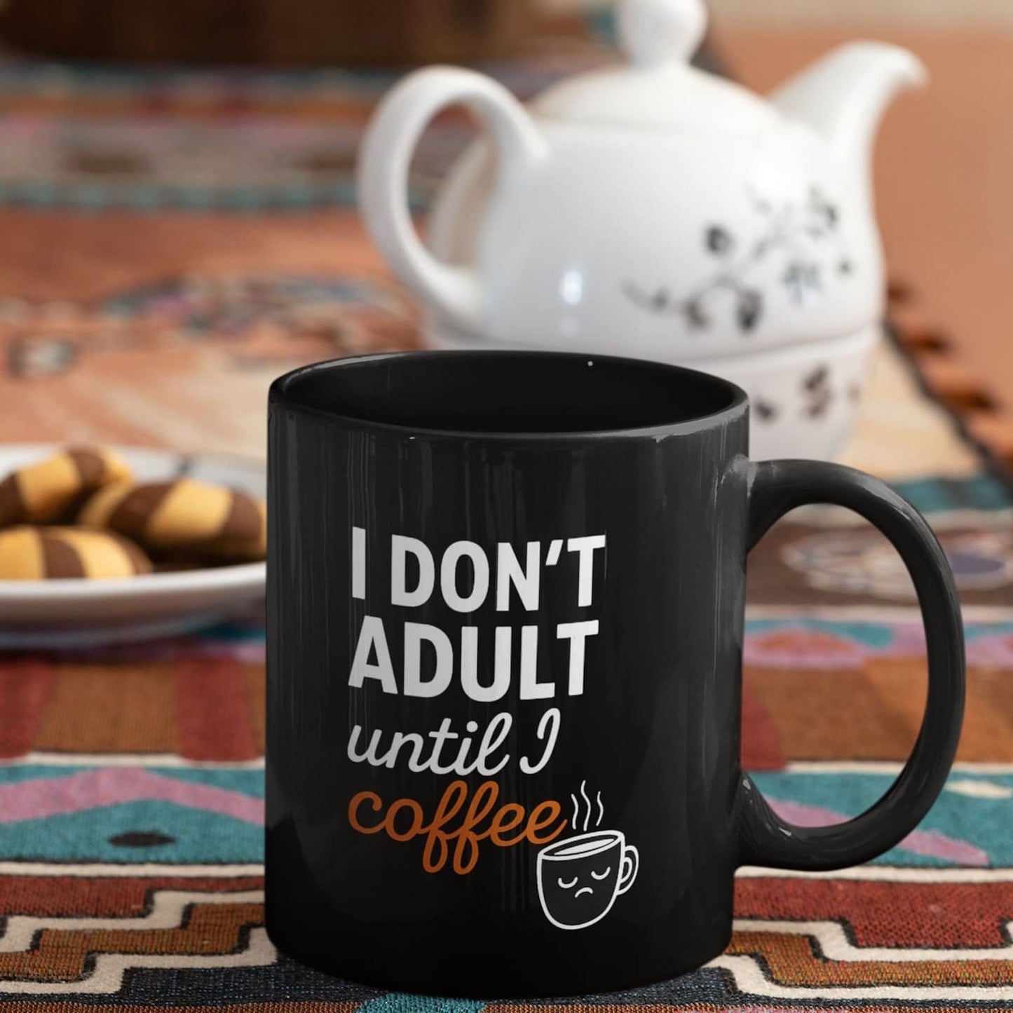 Black ceramic mug with “I don’t adult until I coffee” quote, styled on a colorful tablecloth beside cookies and a white teapot.