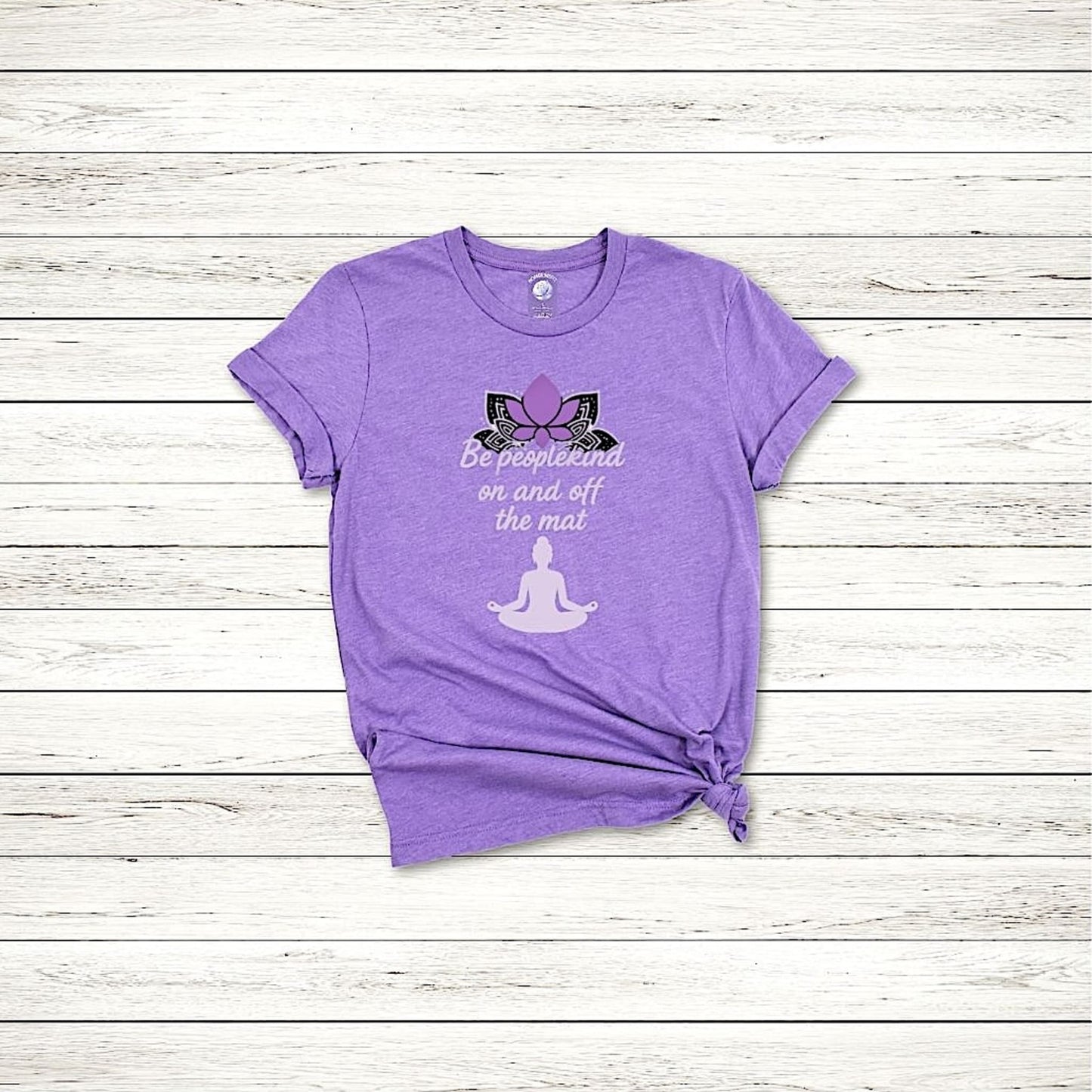 Flat lay of Heather Team Purple Bella Canvas 3001 shirt with yoga quote “Be peoplekind” on wooden background.