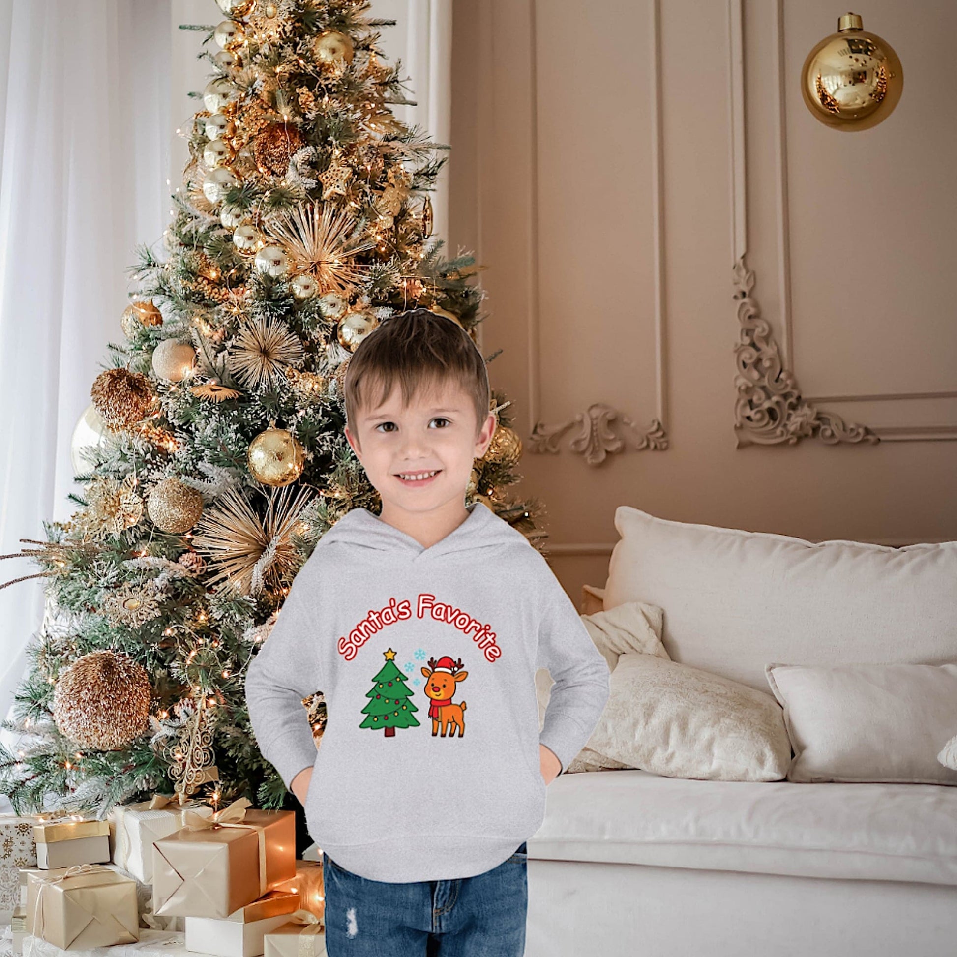 Happy child wearing a gray Santa's Favorite reindeer Christmas hoodie standing by a decorated Christmas tree with gold ornaments and gifts.