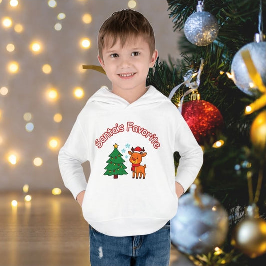 Child wearing a white Christmas hoodie with "Santa's Favorite" text, reindeer and tree design, standing by a decorated Christmas tree.