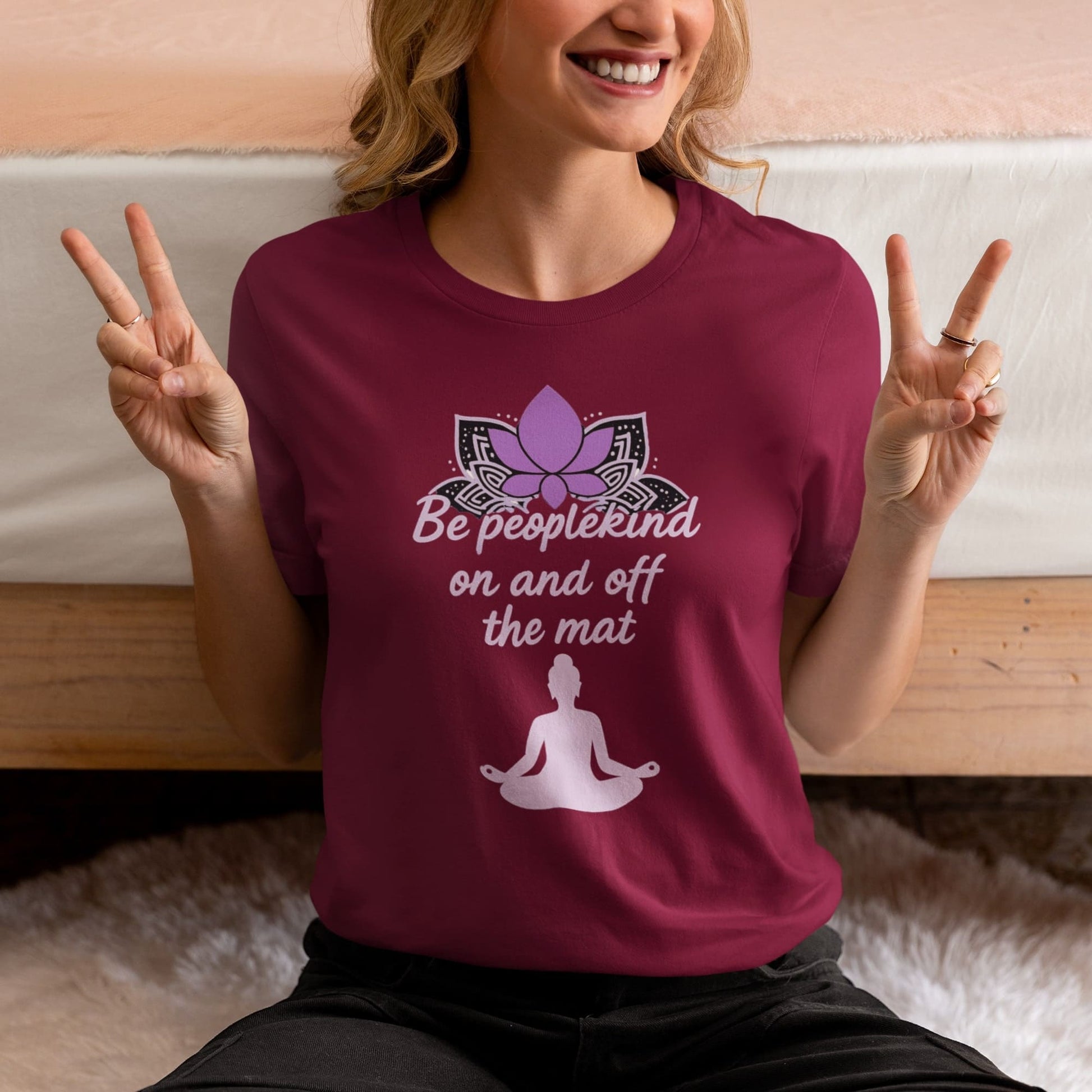 Smiling woman wearing a maroon yoga shirt with lotus flower and meditation quote 'Be peoplekind on and off the mat' while making peace signs