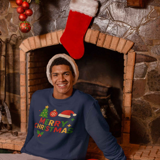 Smiling man in a navy Merry Christmas sweatshirt with festive tree, Santa hat, and holiday decorations sitting by a fireplace.