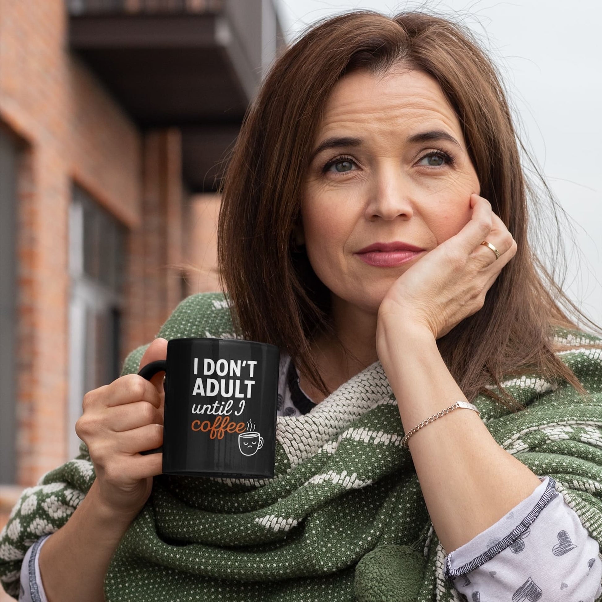 Middle-aged woman holding black coffee mug with “I don’t adult until I coffee” quote, wrapped in green blanket outdoors.
